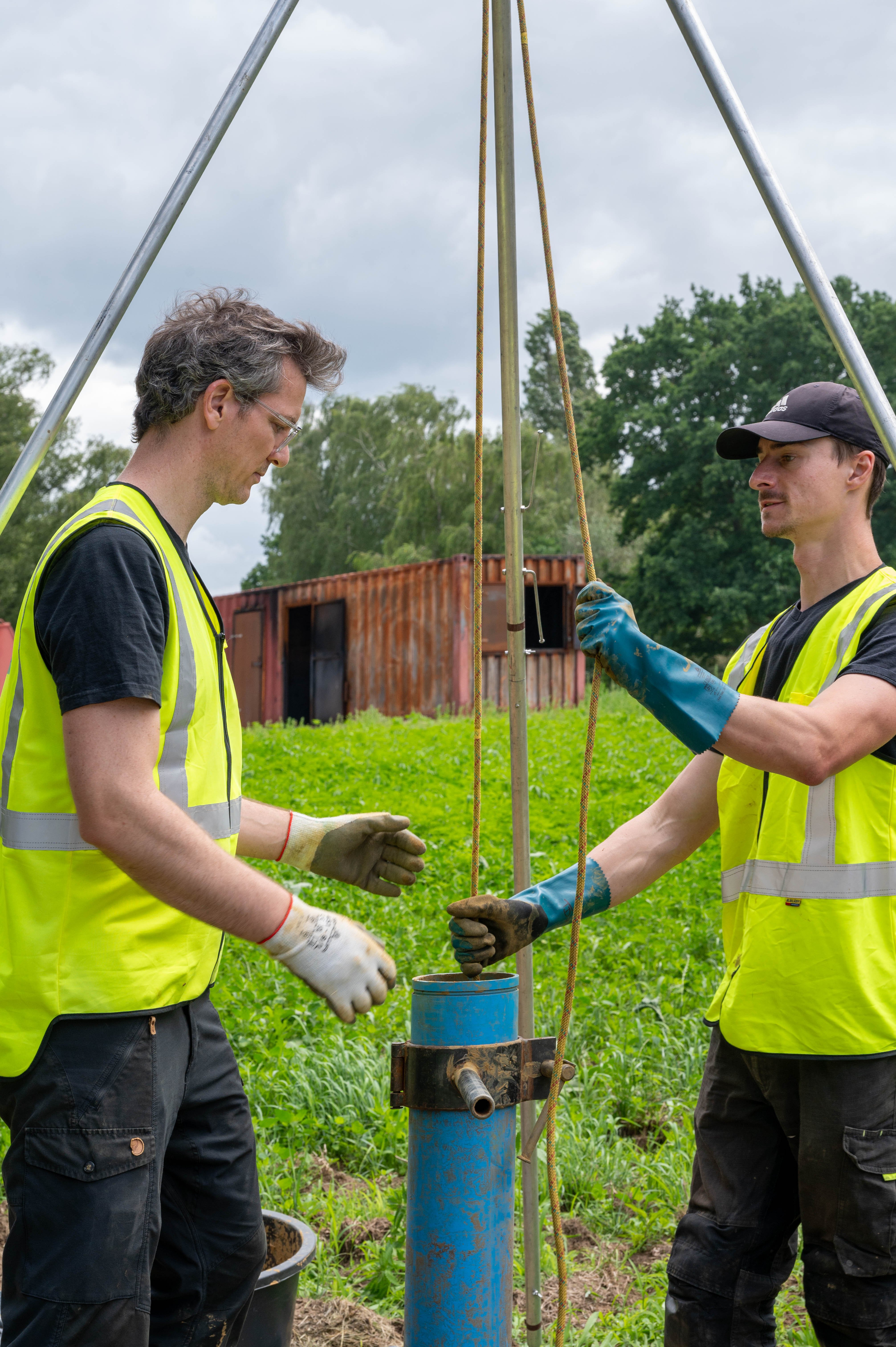 Two iFLUX field technicians in hi-vis vests installing a blue piezometer tube into the ground at Campus Vesta, the casing that will house groundwater flux sensors.